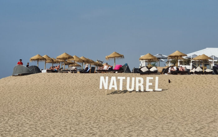 Op het strand van Scheveningen staan grote witte letters van NATUREL. De lucht is blauw en op de achtergrond zie je ligplekken met houten parasols