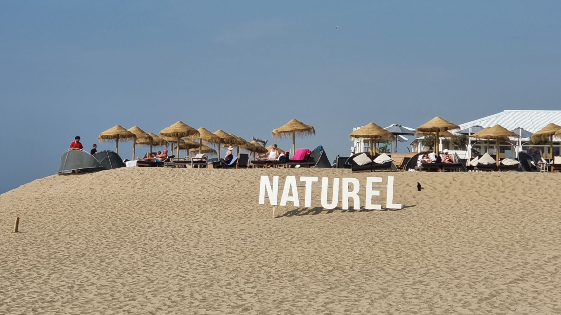 Op het strand van Scheveningen staan grote witte letters van NATUREL. De lucht is blauw en op de achtergrond zie je ligplekken met houten parasols
